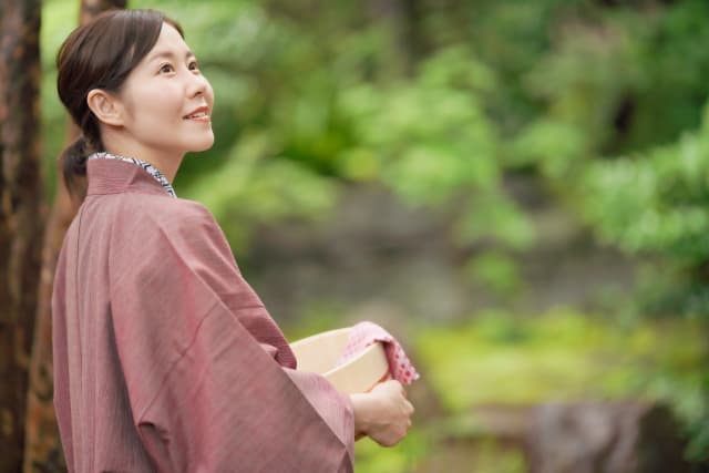 浴衣の女性が木製の湯桶を手に、緑の庭園を背景に微笑む様子